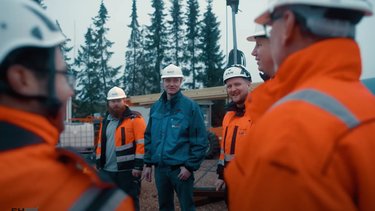 Group of construction workers in orange safety jackets and helmets standing outside on a work site, with trees in the background.