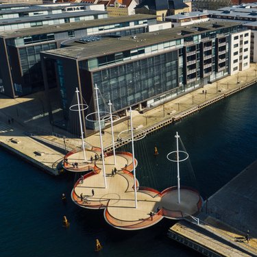 Aerial view of a uniquely shaped pedestrian bridge with circular platforms and tall poles, spanning a canal next to modern buildings.