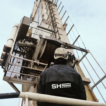 Worker in SH Group jacket looking up at a tall industrial structure with pulleys, cables, and a metal ladder against a cloudy sky.
