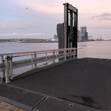 A ferry ramp at a port, extending toward the calm water under a pink and blue sky, with buildings visible across the water.