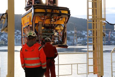 Two workers in safety gear observe a ROV suspended above a waterfront, with a town and cranes visible in the background.