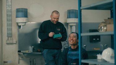 Two workers in a break room, one laughing while seated, and the other smiling and writing on a notepad.