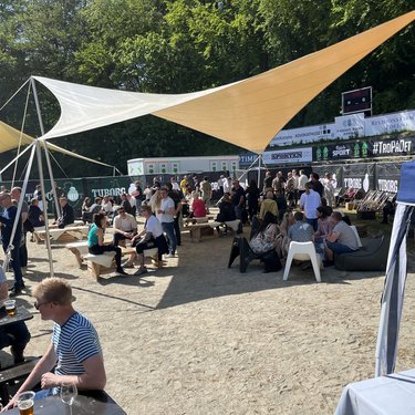 People socializing under large triangular shade tent at an outdoor event, with trees in the background and seated areas.