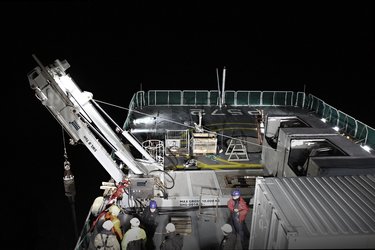 A group of workers wearing helmets operates a crane on a ship's deck at night, with various equipment and a helipad visible in the background.