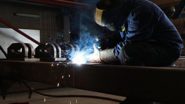 A welder wearing protective gear works on metal, with bright sparks and smoke visible in an industrial setting.
