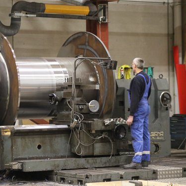A person in blue overalls operates a large industrial metal lathe in a workshop, focusing on a rotating cylindrical workpiece.