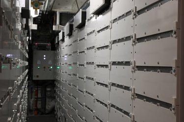 A narrow corridor lined with white server racks, featuring visible wiring and control panels with green lights in a data center environment.