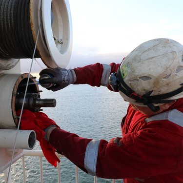 Worker in red safety gear and helmet repairing a winch on a ship's deck, with ocean and coastline in the background.