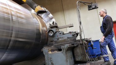 Man operating a large metalworking lathe, focusing on a spinning cylindrical piece in an industrial workshop setting.