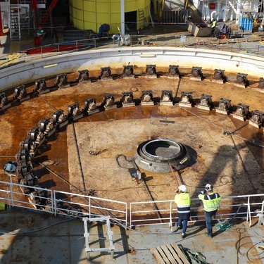 Two workers in safety vests stand near a large circular industrial mechanism on a ship deck, surrounded by machinery and equipment.