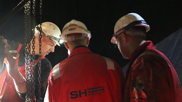 Three workers in red uniforms and helmets collaborate under lighting, with chains visible overhead.