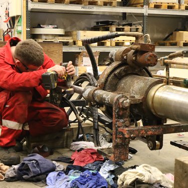 A worker in a red jumpsuit repairs industrial machinery in a workshop, surrounded by tools and cloths.