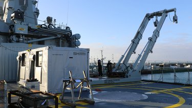 A ship deck featuring a a-frame, equipment containers, tools, and two workers. A large structure and a distant bridge are visible under a clear sky.