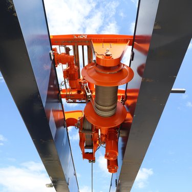 Close-up view of an industrial overhead crane hoist mechanism with steel beams against a blue sky background.