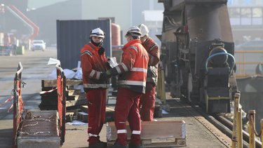 Workers in orange safety gear stand and converse near a crane on an industrial site, with equipment and shipping containers in the background.