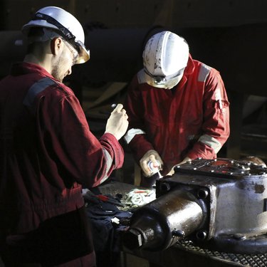 Two workers in red coveralls perform maintenance on heavy machinery under bright light, wearing helmets and safety glasses in an industrial setting.