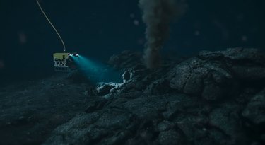 Underwater scene with a robotic vehicle shining a light on a rocky seabed near a vent emitting smoke or gas clouds.