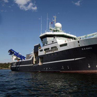 Large research vessel with a blue hull and white superstructure, equipped with cranes and radar, cruising in calm waters under a clear blue sky.