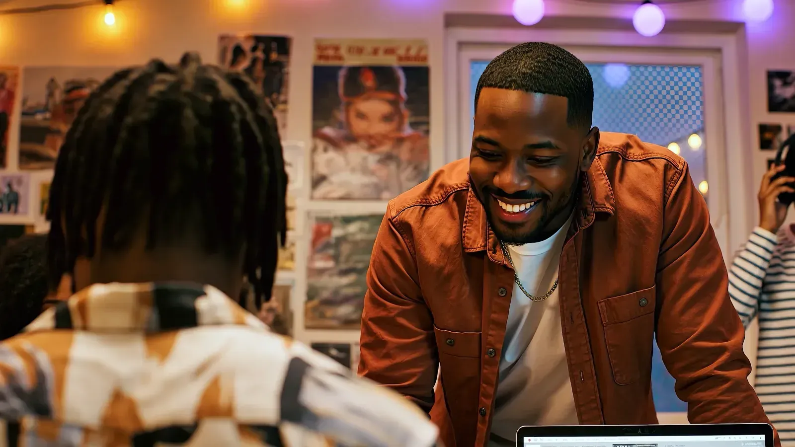 A smiling man in a rust jacket leaning over a desk chatting in a poster-filled studio