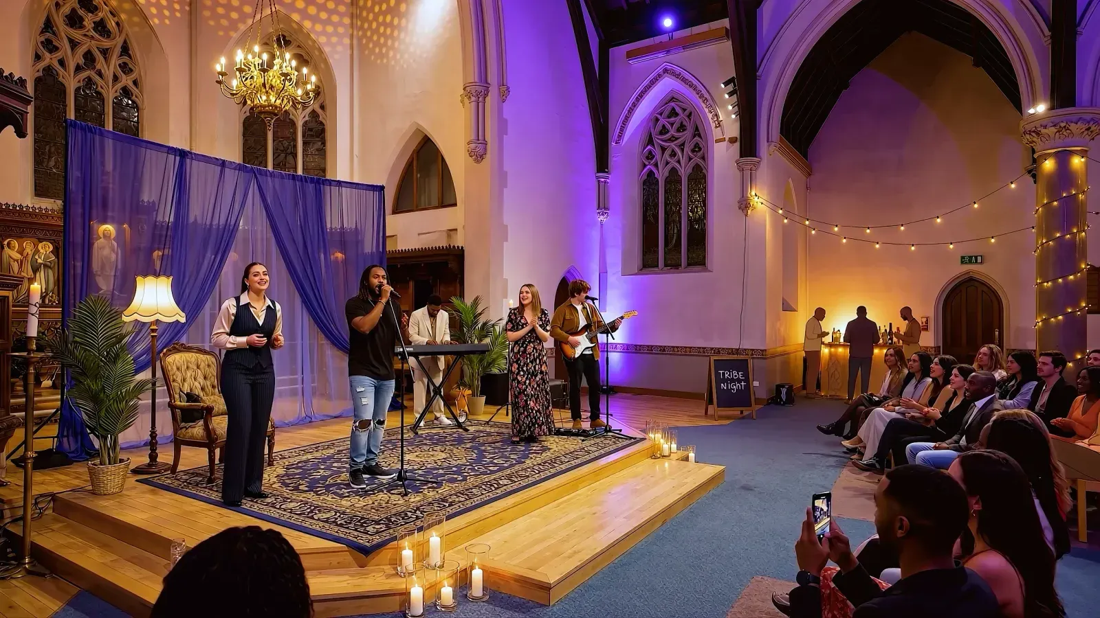 A band performing on a candlelit church stage under a chandelier, a 'Tribe Night' chalkboard beside them