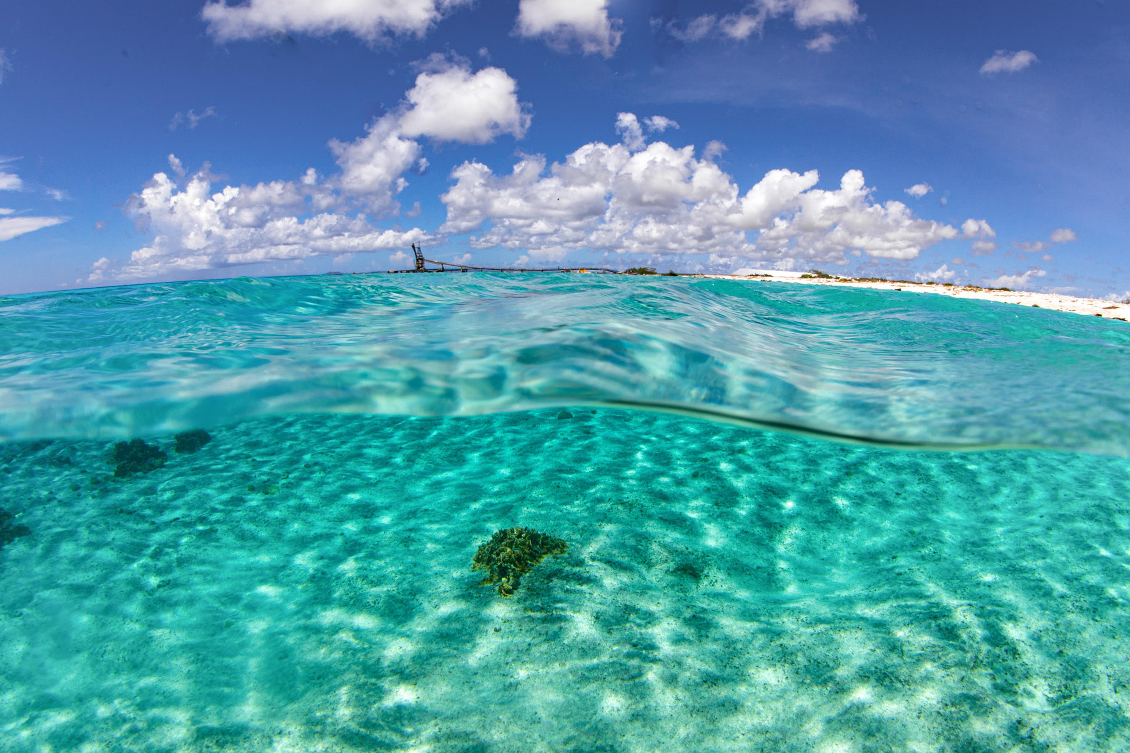 Onderwaterfoto van de blauwe zee en het strand van Bonaire, met een zoutpier op de achtergrond.