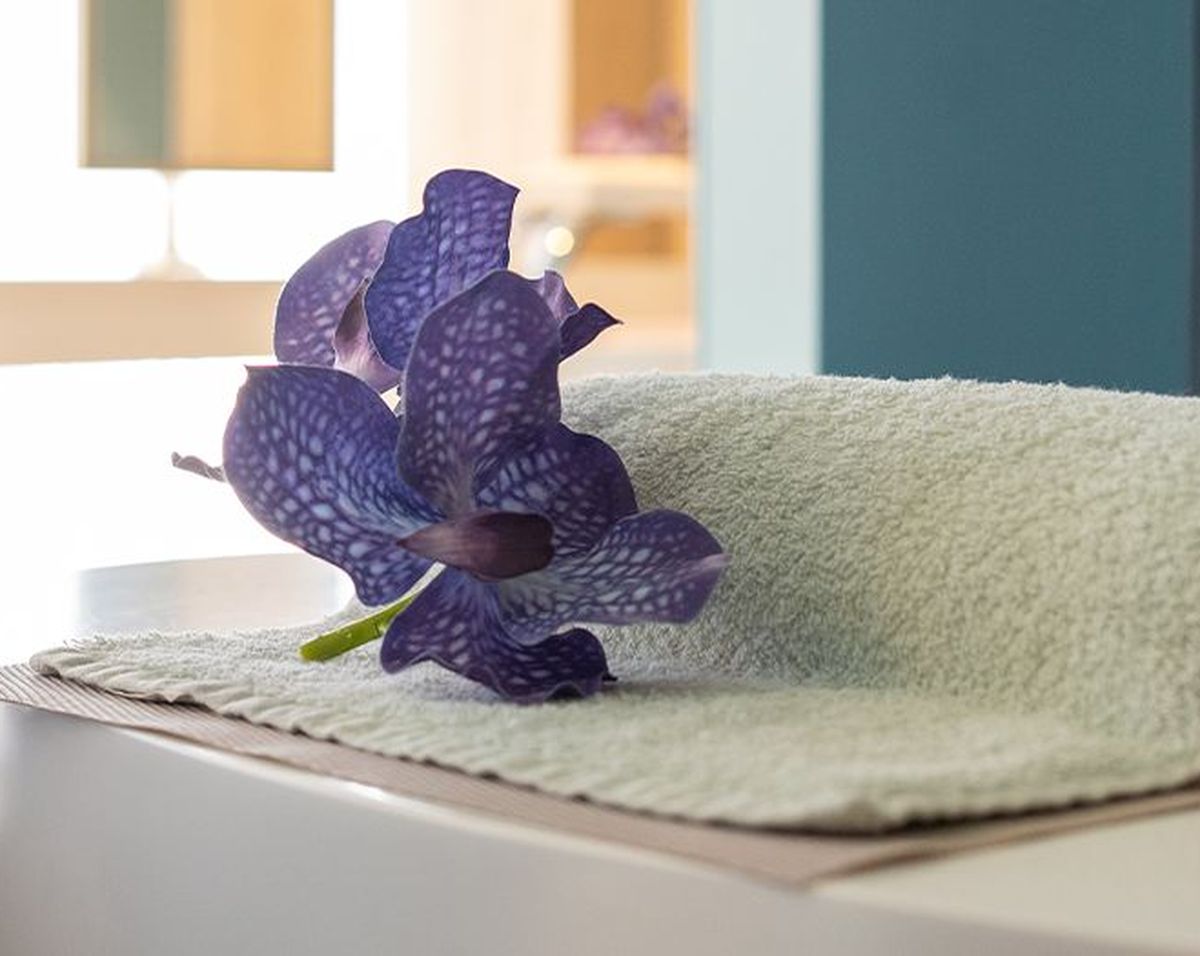 A purple flower rests on a folded towel atop a white surface, with a modern chair and soft lighting in the background.