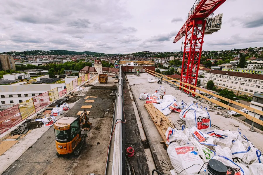 Dachbaustelle mit orangefarbenem Bagger, rotem Baukran und weißen Materialsäcken; im Hintergrund eine Stadt unter bewölktem Himmel.