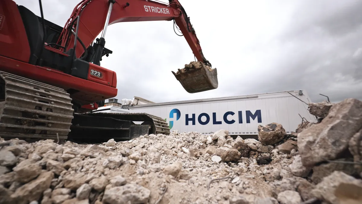 A red STRICKER excavator with a full bucket of rubble in the foreground. A white Holcim trailer is in the background under a cloudy sky.