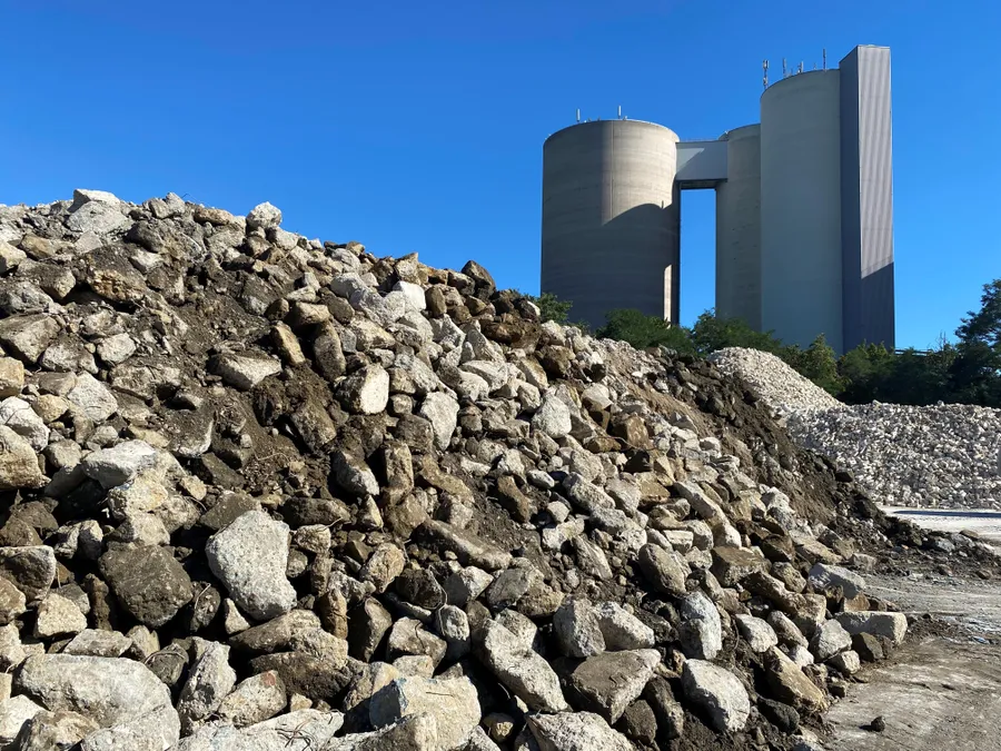 A huge pile of concrete and dirt rubble in the foreground, with two massive gray industrial silos connected by a bridge against a clear blue sky.
