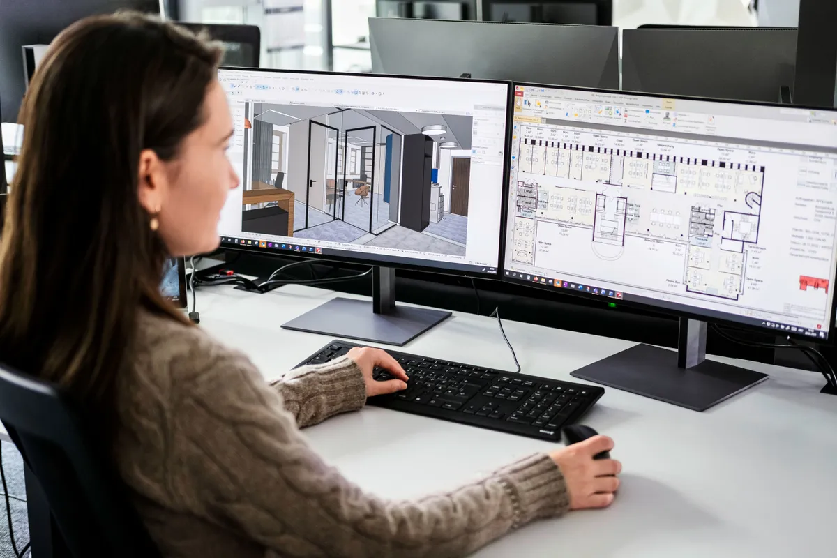 An architect working at two monitors displaying construction drawings and 3D models.