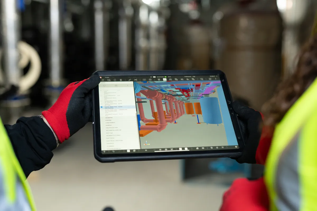 Two people in safety vests are using a tablet to view a 3D model of industrial pipes and tanks.