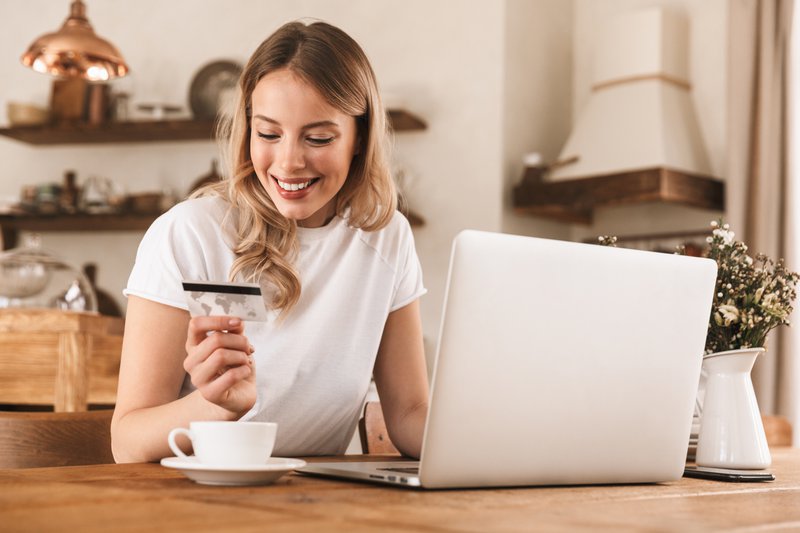 Porträt einer schönen blonden Frau in den 20ern, die ein lässiges T-Shirt trägt und einen Laptop und eine Kreditkarte zum Online-Shopping benutzt, während sie in einem gemütlichen Café sitzt