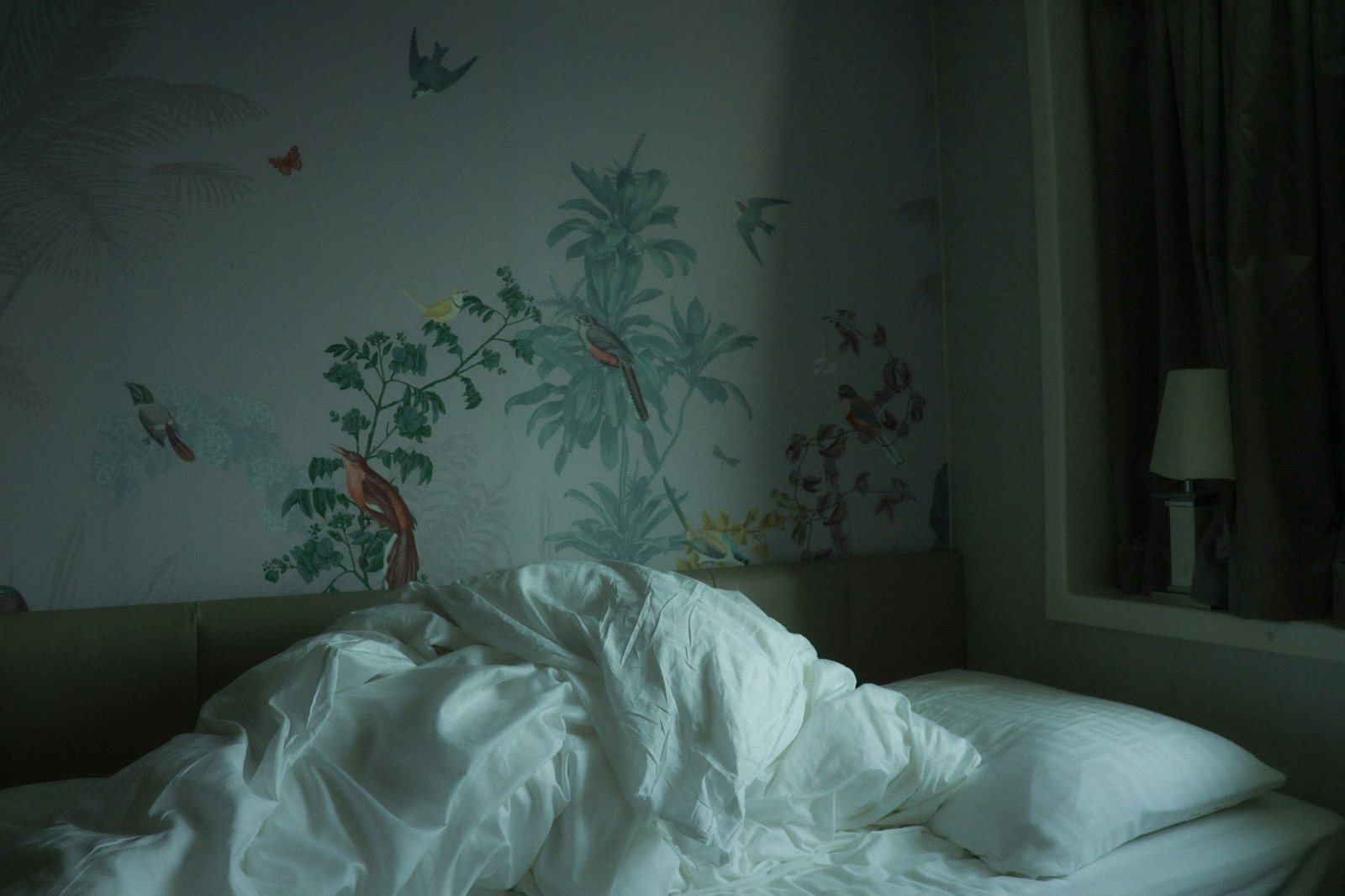 An unmade white bed beneath hand-painted botanical wallpaper.
