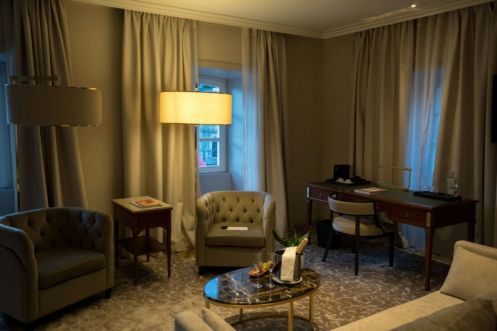 A warmly lit suite sitting room with curtains, armchairs and a writing desk.