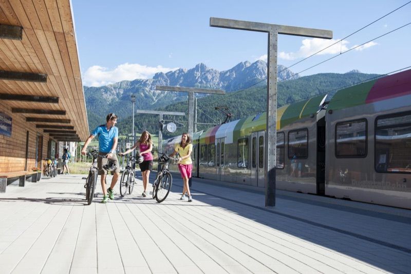 Drei Fahrgäste schieben ihre Räder über einen Bahnsteig zu einem wartenden Zug, dahinter die Berge