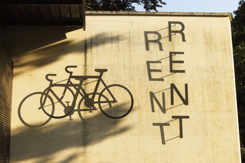 Ein Fahrradsymbol und der Schriftzug „RENT“ an einer sonnenbeschienenen Betonwand