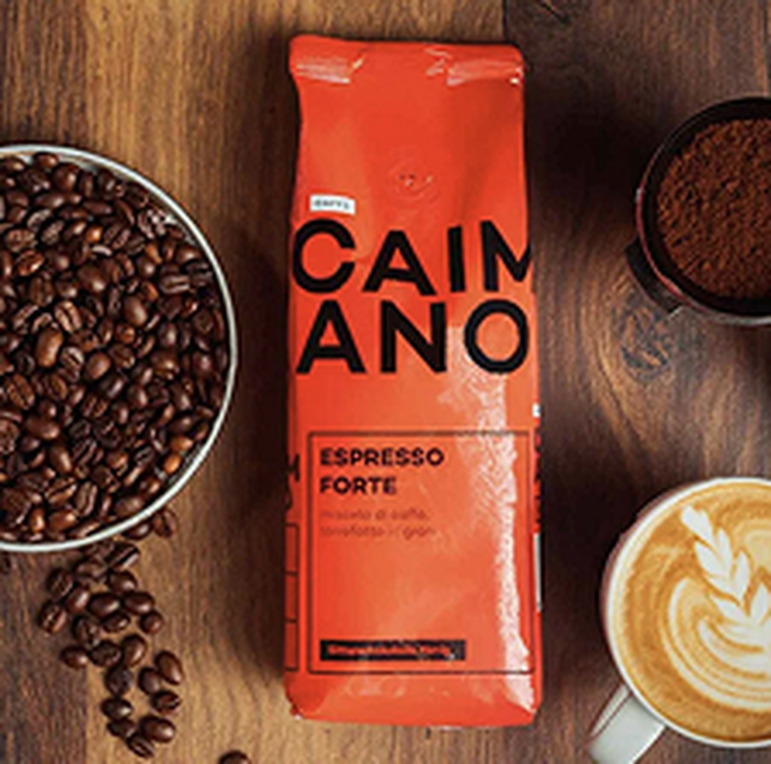 Bag of Caimano espresso forte on a wooden table with a bowl of coffee beans, ground coffee, and a cup of latte with latte art.