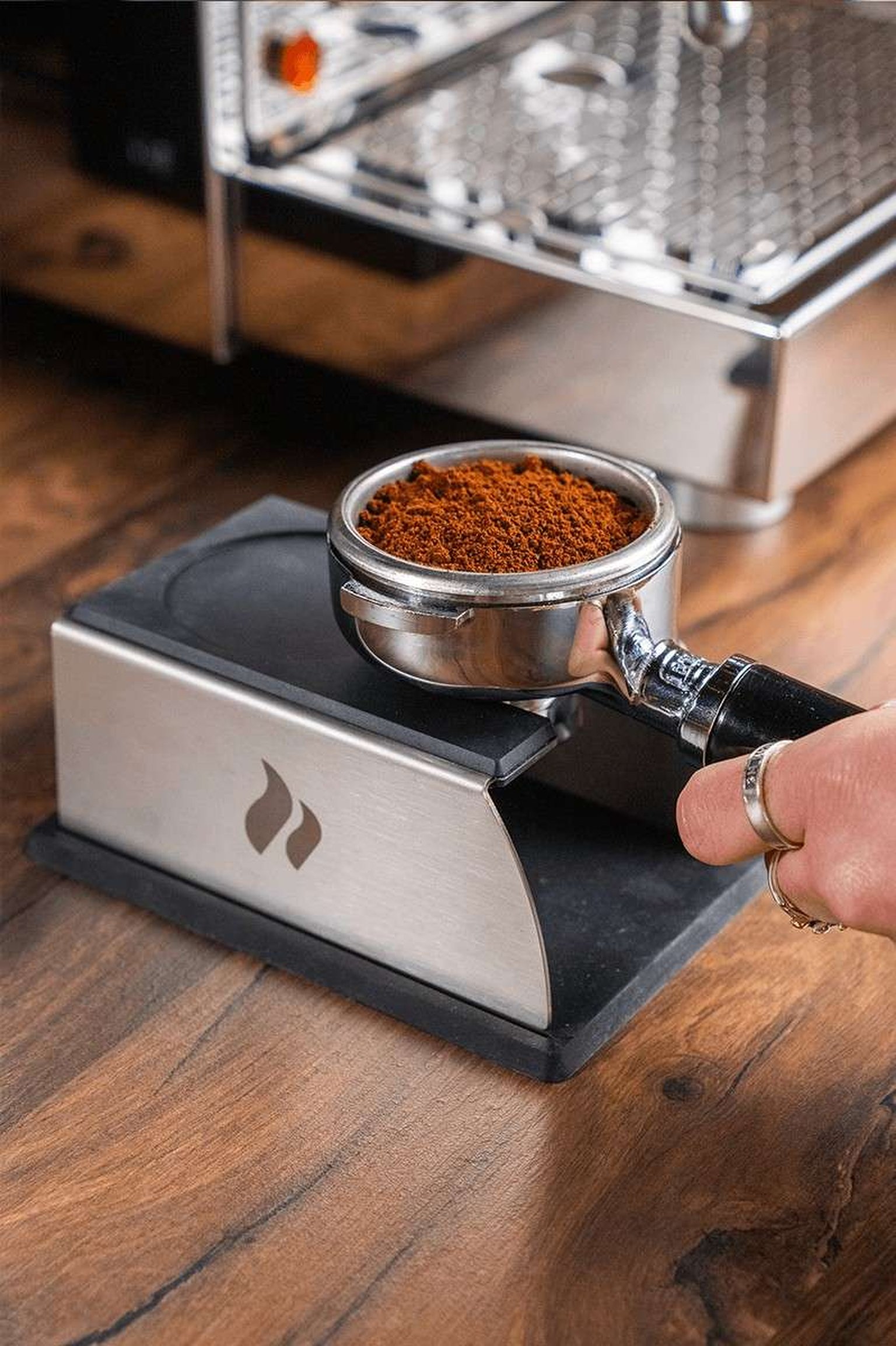 A hand uses a tamper on coffee grounds in a portafilter on a wooden countertop.