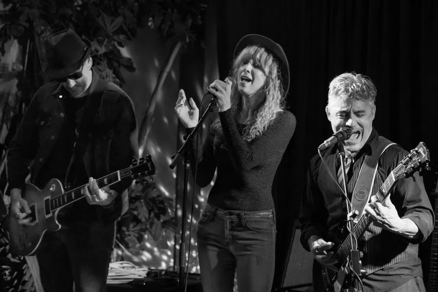 Zwart-witfoto van drie zangers en gitaristen midden in een nummer, met gespikkeld licht op het gordijn