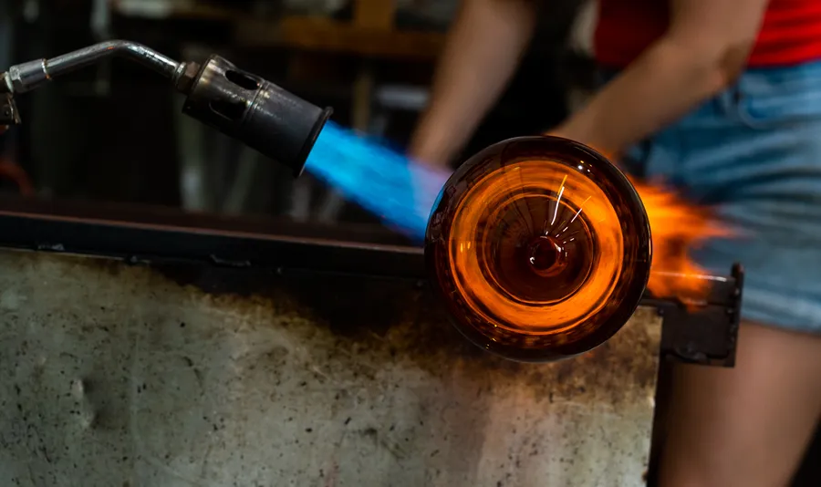 Een brander met blauwe vlam verhit een gloeiend amberkleurig glazen object in een glasstudio