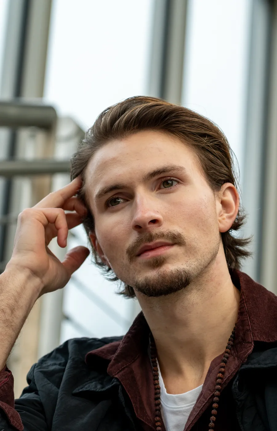 Jonge man met achterovergekamd haar poseert bedachtzaam, met glazen architectuur achter hem