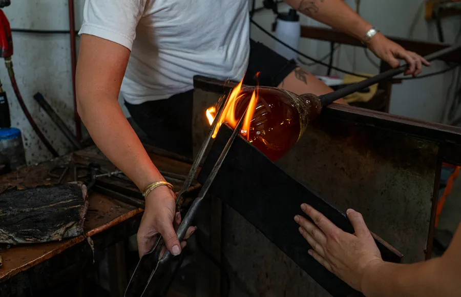 Twee glasblazers vormen gesmolten amberkleurig glas in een werkplaats met opstijgende vlammen