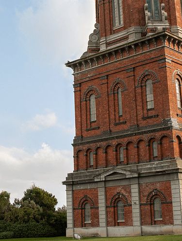 Two people walking along a path next to the historic Invercargill Water Tower, a tall red-brick structure with a domed top, set against a clear blue sky with scattered clouds.