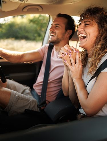Happy couple laughing inside a car on a road trip, man driving while woman smiles with hands clasped, green trees in background.