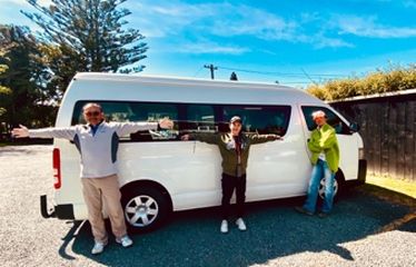 Three people standing beside a white van, arms wide open, on a sunny day