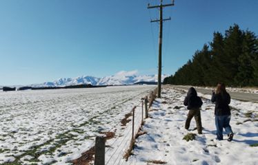 Two people walking on a snowy roadside with mountains in the background