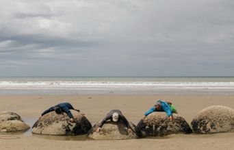 Three people lying on large round boulders at the beach under a cloudy sky