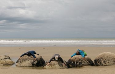 Three people lying on large round boulders at the beach under a cloudy sky