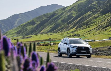White Toyota hybrid SUV driving along a scenic road with green hills and purple flowers