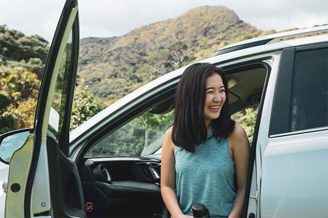 Image of woman laughing, sitting on the front seat of a car parked in front of dense bush